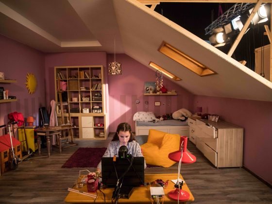 Mergaitė sėdi priešais kompiuterį filmavimo erdvėje jai sukurtame vaikų kambaryje