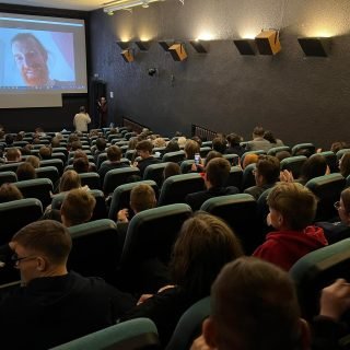 Kino salė pilna žiūrovų, ekrane matomas Zoom prisijungęs režisierius