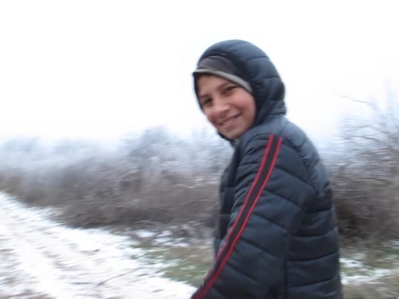 Laukai ir kelias, užkloti snieku. Priekyje stovi atsisukęs besišypsantis paauglys su kapišonu.
